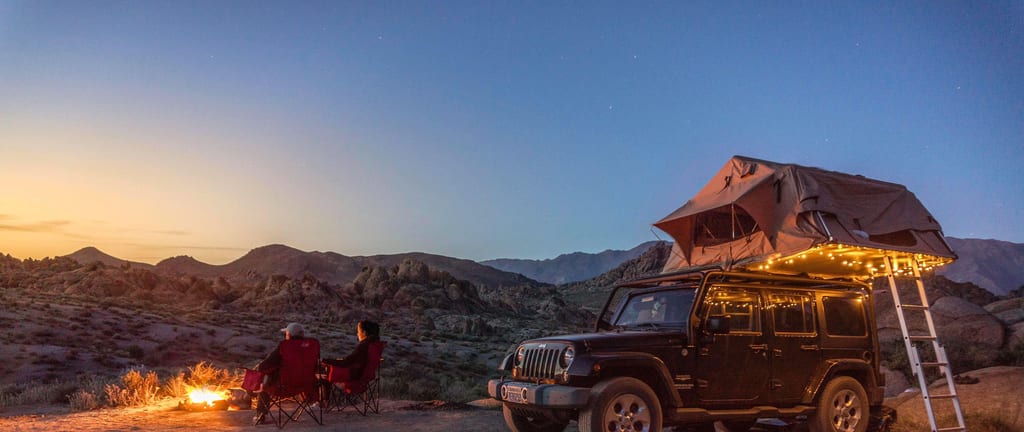 Desert camping scene with Jeep, roof tent, campfire, and two people at dusk in rocky landscape.