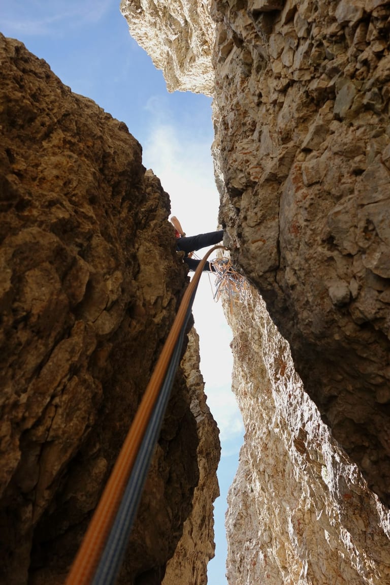 Rock climber ascending narrow canyon with rope and gear between towering stone walls.